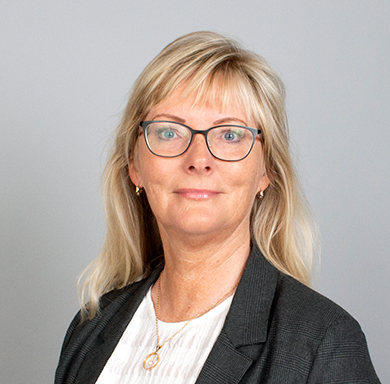 Supply Chain Manager at TAGARNO, Helle Anthonsen-Rahbek, in white blouse and grey blazer, cropped at the chest, smiling at the camera in front of a grey wall, classic employee photo