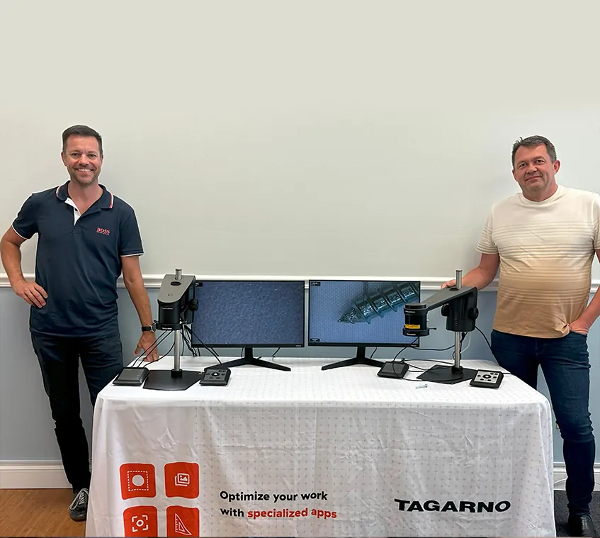 Two employees at TAGARNO's office in Tampa, Florida, on either side of a table that's covered in a white table cloth and with 2 TAGARNO digital microscopes on. The two men are smiling at the camera.